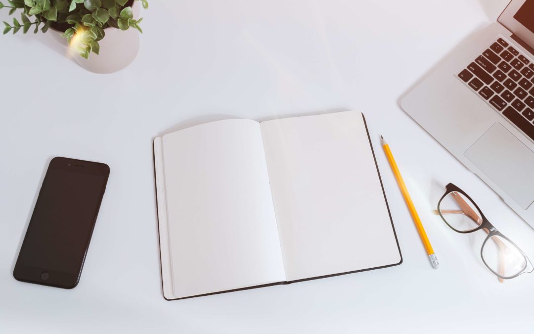 Notebook on a table with pencil, glasses, computer, and phone - tools to help you get your writing project done